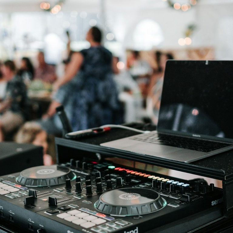 DJ equipment in focus with a lively crowd and a performer in the background.